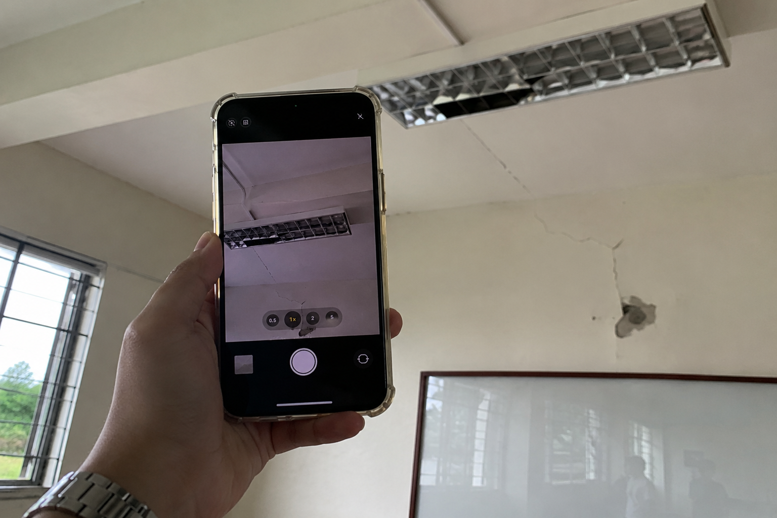 A hand holding a phone and photographing a cracked ceiling and light fixture in a classroom.